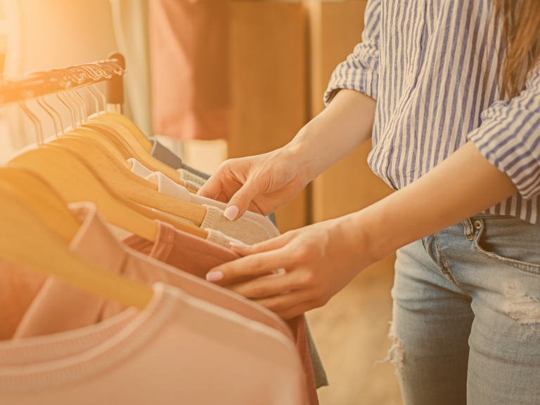 Junge Frau in Jeans sieht Kleiderständer mit nachhaltigen Produkten im Ladengeschäft durch