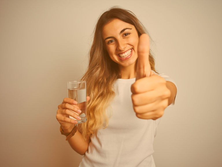 Junges, langhaariges Mädchen hält in ihrer einen Hand ein Glas Wasser und macht mit der anderen Hand das Daumen-hoch-Zeichen 