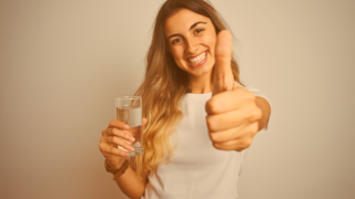 Junges, langhaariges Mädchen hält in ihrer einen Hand ein Glas Wasser und macht mit der anderen Hand das Daumen-hoch-Zeichen 