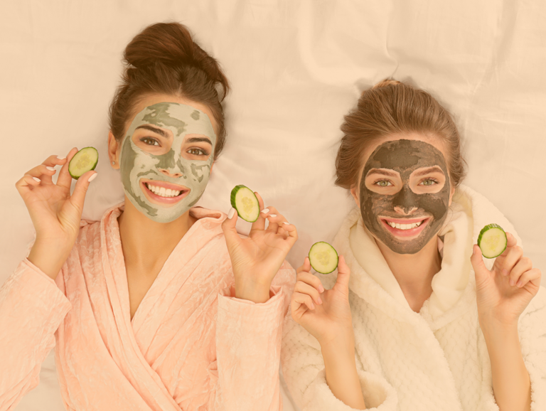 Zwei Freundinnen liegen im Bademantel und grüner Gesichtsmaske im Bett und halten je beidhändig Gurkenscheiben in den Händen