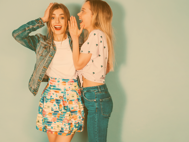 Zwei blonde Jugendliche stehen zusammen und die eine tuschelt der anderen mit überraschtem Gesichtsausdruck etwas ins Ohr