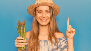 Jugendliche mit Strohhut hält in der rechen Hand eine Bund frischen, grünen Spargel und der linke Zeigefinger ist gehoben 