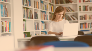 Jugendliche sitzt in einem hellen Raum vor Bücherregalen schreibend am Laptop, um sie herum liegen Unterlagen auf dem Tisch