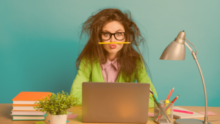 Schülerin mit zerzaustem Haar und Stift zw. Oberlippe und Nase geklemmt, sitz am Schreibtisch hinter dem Laptop